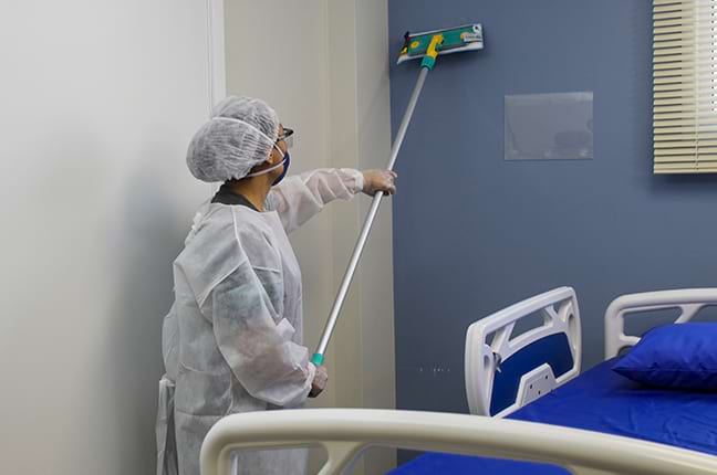 Especialista em desinfecção hospitalar usando MOP para limpar parede de quarto hospitalar.