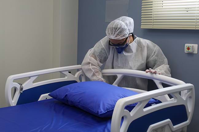 Profissional de saúde limpando uma cama hospitalar com equipamento de proteção.
